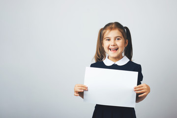 Naklejka premium Little funny girl in a school uniform show an empty paper. Copy space. The concept of education, back to school, etc.