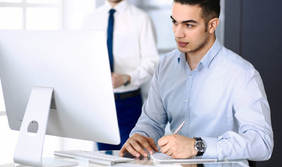 Businessman working with computer in modern office, male colleague at background . Headshot of arab male entrepreneur or manager at workplace. Business concept, teamwork and multi ethnic