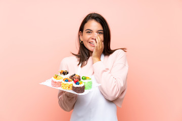 Young girl holding lots of different mini cakes over isolated background with surprise facial expression
