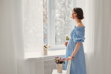 Beautiful pregnant young girl near the window, behind which winter and snow . The concept of family values, warmth and home comfort