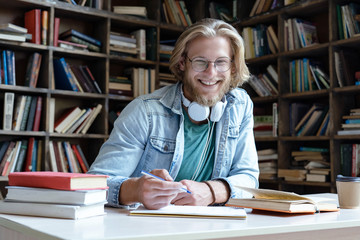 Happy guy university student in library learning with books headphones doing work assignment looking at camera sit at desk, smiling young man teacher wear glasses study write notes, portrait