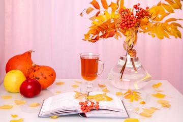 Still life with autumnal colorful red rowan branch with foliage in a glass vase, pumpkins, apples and open book