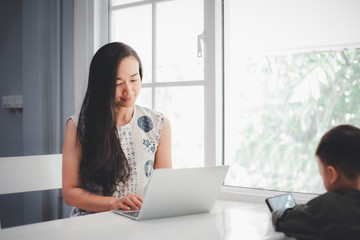 Beautiful Asian girls using laptop computer and her son looking at smartphone. Raise children and work concept.