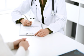 Woman doctor and patient sitting and talking at medical examination at hospital office, close-up. Therapist filling up medication history records. Medicine and healthcare concept