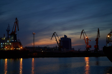 Fototapeta premium Colorful sunset over sea port and industrial cranes. Bright sunset in seaport. Large silhouettes of cargo cranes. Beautiful landscape with fiery sunset sky and sea.