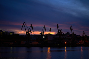 Fototapeta na wymiar Colorful sunset over sea port and industrial cranes. Bright sunset in seaport. Large silhouettes of cargo cranes. Beautiful landscape with fiery sunset sky and sea.
