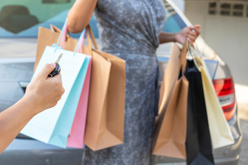The hand of a woman holding a car key and back of a woman carrying a shopping bag with the car in the background. They returned from shopping.buy,sell,Photo concept Lifestyle and Shopping.
