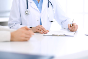 Woman doctor and patient sitting and talking at medical examination at hospital office, close-up. Physician filling up medication history records. Medicine and healthcare concept
