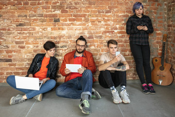 Fototapeta premium Group of happy caucasian young people behind the brick wall. Sharing a news, photos or videos from smartphones, laptops or tablets, playing games and having fun. Social media, modern technologies.