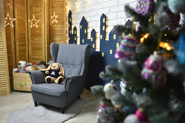 Interior of a children's playroom decorated for the New Year holiday, with a Christmas tree, an armchair and soft toys, against a white brick wall.