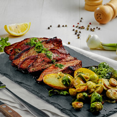 Delicious pan-seared steak with vegetables on a stone board. Meat meal with brussels sprouts, potatoes, onions and herb butter on a white wooden table.
