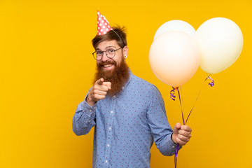 Redhead man with long beard holding balloons over isolated yellow background points finger at you...