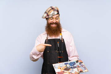 Redhead artist man with long beard holding a palette over isolated blue background and pointing it