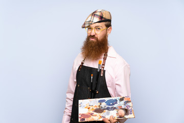 Redhead artist man with long beard holding a palette over isolated blue background with sad...