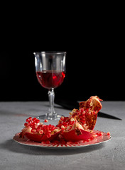 Open pomegranate fruit on a plate, on the background of pomegranate juice in a glass on a long stem and knife on a black background.
