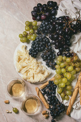 Swiss cheese tete de moine on ceramic board, black and green grapes and two glasses of white wine, served with bread sticks on pink marble background. Flat lay, space