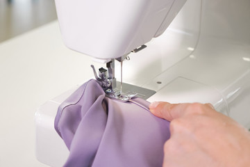 Processing tissue of fabric edge. Seamstress working at sewing machine making stitches seams with straight stitch on purple cloth, hands closeup. Clothing tailoring business manufacture.