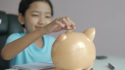 Little Asian girl puttinwg the coin into piggy bank and smile with happiness for money saving to wealthness in the future of education concept select focus shallow depth of field