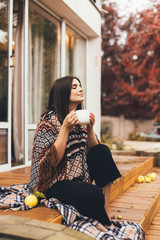 Young woman relaxing with hot tea on cozy terrace of wooden countryhouse. Autumn cozy day.