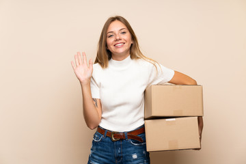 Young blonde woman over isolated background holding a box to move it to another site and saluting