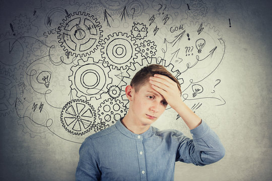 Desperate teen boy covering his forehead with hand. Hopeless situation, hard thinking as cogwheel gear brain arrows and mess thoughts over grey wall. Concept of mental, psychological trauma.