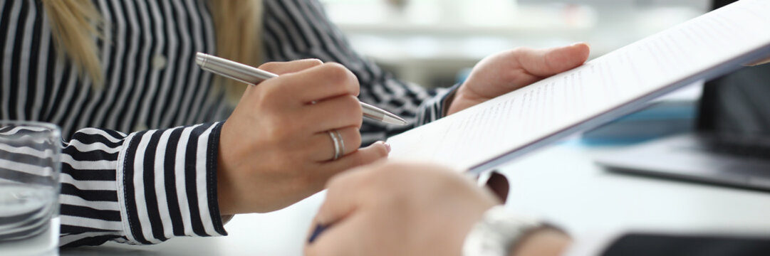 Focus On Tender Female Hand Signing Corporate Contract. Woman In Trendy Clothes Holding Distinctive Document And Sitting In Modern Biz Office With Partner. Blurred Background