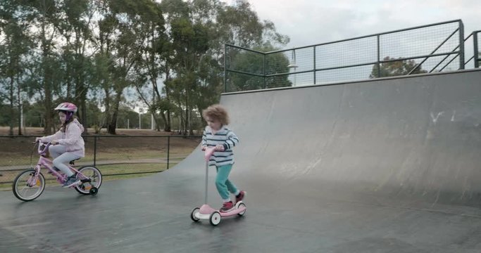 a small boy skates on a scooter and girl rides a bicycle on a skateboard ramp half pipe circling around between the ramps