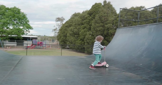 a small boy rides a pink three wheeled scooter on a skateboard half pipe ramp, skating from side to side
