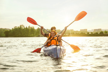 Happy young caucasian couple kayaking on river with sunset in the backgrounds. Having fun in leisure activity. Happy male and female model laughting on the kayak. Sport, relations concept. Colorful.