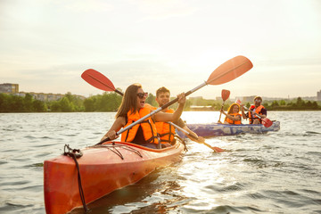 Happy young caucasian group of friends kayaking on river with sunset in the backgrounds. Having fun in leisure activity. Happy male and female model laughting on the kayak. Sport, relations concept. - obrazy, fototapety, plakaty