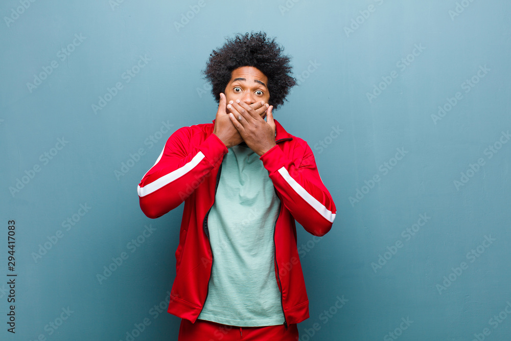 Wall mural young black sports man covering mouth with hands with a shocked, surprised expression, keeping a secret or saying oops against grunge wall - Wall murals