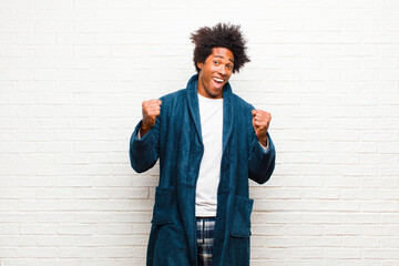 young black man wearing pajamas with gown feeling shocked, excited and happy, laughing and...