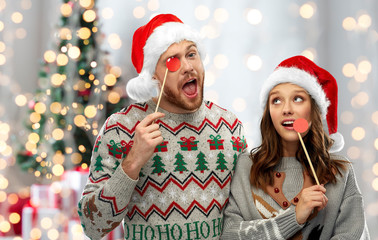 christmas, photo booth and holidays concept - happy couple in ugly sweaters posing with party props over festive lights background