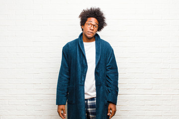 young black man wearing pajamas with gown looking puzzled and confused, biting lip with a nervous...
