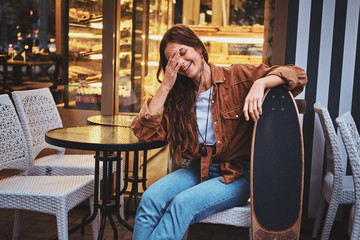 Happy smiling woman is sitting at cafe outside while holding her longboard.