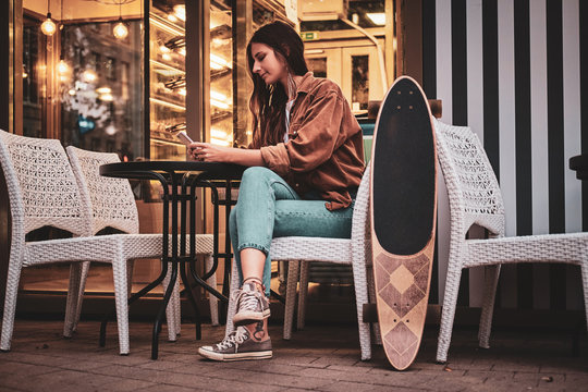 Pretty woman is sitting at cafe outside with her longboard while chatting by mobile phone. - Powered by Adobe