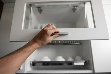 Hand holds the door of the dryer with utensils. Dish drying metal rack with white clean plates. Traditional comfortable kitchen. Open white dish draining closet with wet dishes of plates, bowls.