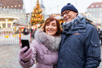 technology, winter holidays and people concept - happy senior couple taking selfie by smartphone at christmas market on town hall square in tallinn, estonia