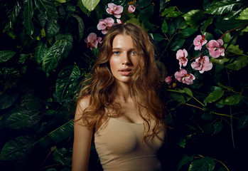 A beautiful blue-eyed long-haired brown-haired woman stands on a background of exotic plants and pink orchids.