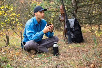 caucasian male hunter resting in forest. drinking tea from thermos. shotgun, ammunition, backpack...