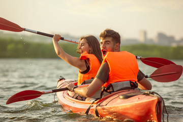 Happy young caucasian couple kayaking on river with sunset in the backgrounds. Having fun in leisure activity. Happy male and female model laughting on the kayak. Sport, relations concept. Colorful.