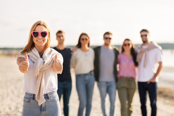 Obraz premium friendship, valentine's day and people concept - happy woman in heart-shaped sunglasses with group of friends on beach in summer showing thumbs up