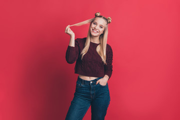 Photo of amazing millennial lady holding hands in pockets positive mood playing with long curl wear knitted pullover and jeans isolated pastel red color background