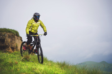 Naklejka na ściany i meble Front view of a man on a mountain bike standing on a rocky terrain and looking down against a gray sky. The concept of a mountain bike and mtb downhill