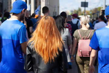 Girls with red hair walks around the city in a crowd of people. A lot of people are walking down the street. Bright girl goes with the guys.