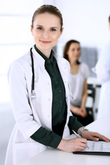 Doctor woman at work with patient and colleague at background. Physician filling up medical documents or prescription while standing in hospital reception desk. Data in medicine and health care