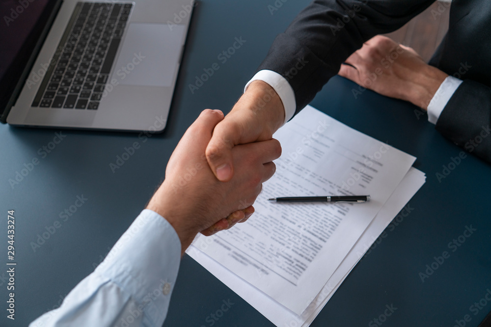 Poster top view of two businessmen shaking hands in blurred office with computer and contract on table. con