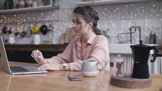 A young woman sitting at the kitchen table using a laptop. Stock footage. Beautiful brunette working at home on her computer with a cup of hot drink standing on the wooden table.