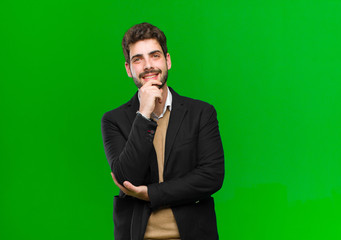 young businessman looking happy and smiling with hand on chin, wondering or asking a question,...