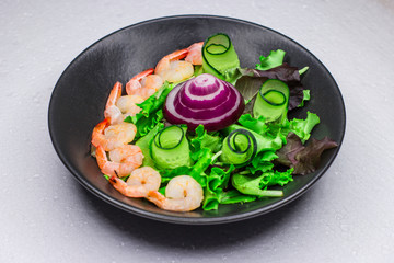 Crimean onion. Green cucumber and arugula. White table. Salad with cabbage leaves and golden shrimps. Black dish. Concept for a tasty and healthy meal. Dark background.
